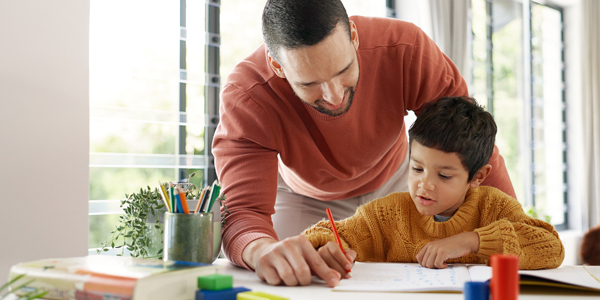 Pai desenvolvendo com o filho atividades de alfabetização, o menino sentado com lápis e caderno e o pai em pé